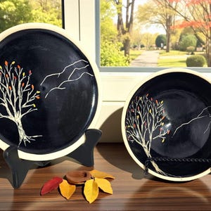 May include: Two black ceramic plates with a white tree design and colorful leaves. One is a plate, the other a bowl. Both have a white rim and are displayed on black stands. Autumn-themed decorative tableware.