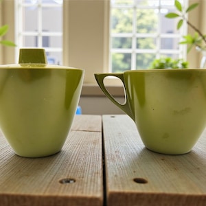 May include: A light green tea cup and sugar bowl set. The sugar bowl has a lid with a square handle. The cup has a triangular handle. Both pieces are on a wooden surface.