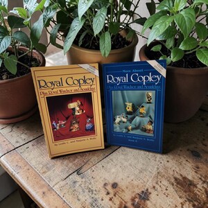 May include: Two books titled "Royal Copley" and "More About Royal Copley" are displayed. The first book has a yellow cover with a picture of figurines. The second book has a blue cover with a picture of figurines. Both books are on a wooden surface.
