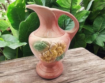 May include: A pink ceramic pitcher with a floral design. The pitcher has a wide mouth and a handle. The flowers are yellow and green.