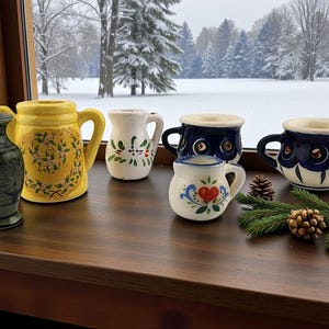 May include: A collection of ceramic pitchers and mugs in various sizes and designs. The color palette includes yellow, white, and dark blue, with floral and heart motifs. The items are arranged on a wooden surface with a snowy outdoor backdrop.