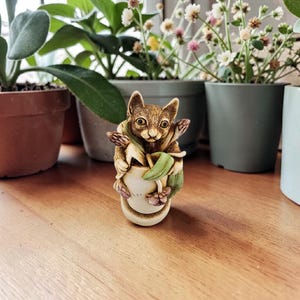 May include: A detailed figurine of a squirrel holding a small pot with green leaves and pink flowers. The squirrel is brown with large, expressive eyes. The pot is off-white with a textured surface. The figurine is set against a backdrop of potted plants.