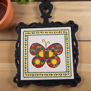 May include: Black cast iron trivet with a decorative butterfly tile insert. The tile features a red butterfly design with yellow and blue accents, set against a white background. The trivet has a square shape with ornate edges and a handle.