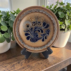 May include: A round ceramic plate featuring a speckled tan glaze with a dark blue embossed butterfly and floral scroll design in the center. The rim has a textured, scalloped pattern, and the plate is displayed on a black plastic stand.