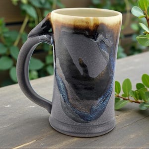 May include: A handmade ceramic mug with a unique handle design. The mug features a dark gray base with flowing patterns of blue, black, and brown. The rim has a light tan color. The mug is sitting on a wooden surface.