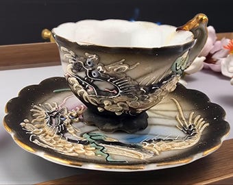May include: An ornate teacup and saucer set. The cup and saucer are black with a detailed dragon design in gold, white, and shades of gray. The cup has a scalloped rim and a gold-accented handle. The saucer has a scalloped edge with gold trim.