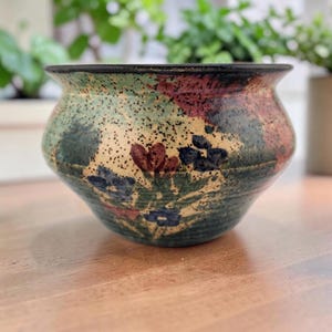 May include: A rounded ceramic bowl featuring a speckled beige base with hand-painted floral motifs in shades of blue, red, and green. The bowl has a wide, flared rim with a dark, solid-colored edge and a smooth, glossy finish throughout the exterior.