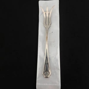 May include: A silver-toned metal cocktail fork with three slender tines and a long, narrow handle. The handle features a decorative geometric cutout pattern near the base. The fork is encased in a clear, rectangular plastic protective sleeve.