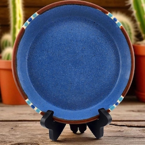 May include: A round, blue ceramic plate with a textured surface and a brown rim. The rim is decorated with a pattern of blue, white, and tan rectangles. The plate is displayed on a black stand, with cacti in the background.