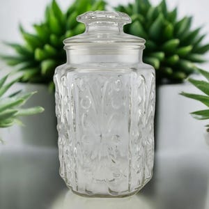 May include: A clear glass jar with a lid, featuring a decorative embossed floral pattern. The jar is empty and has a vintage look.