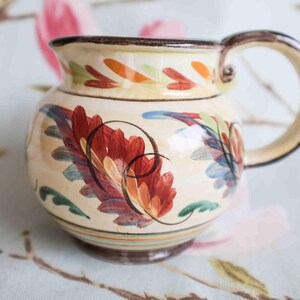 May include: A cream-coloured ceramic jug with a brown handle. The jug is decorated with hand-painted floral designs in shades of red, orange, blue, and green.
