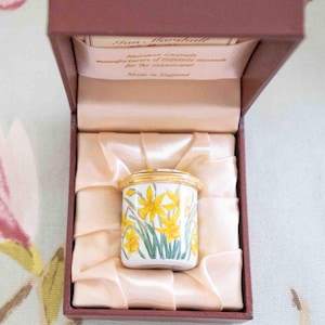 Puede incluir: Una pequeña caja cilíndrica para baratijas con tapa, con un diseño floral de narcisos amarillos y hojas verdes. La caja se presenta dentro de una caja marrón forrada con tela de satén. La tapa y el borde están adornados con oro. La caja está marcada "Made in England".