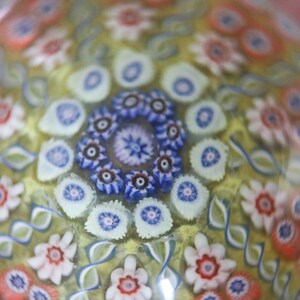 May include: A close-up of a glass paperweight with a colorful, intricate pattern of flowers and swirls. The paperweight is made of clear glass with a yellow background and features a variety of colors, including red, blue, white, and pink.