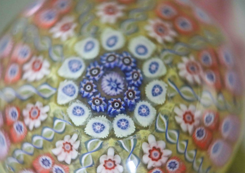 May include: A close-up of a glass paperweight with a colorful, intricate pattern of flowers and swirls. The paperweight is made of clear glass with a yellow background and features a variety of colors, including red, blue, white, and pink.