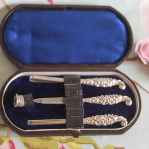 Puede incluir: Un juego de manicura vintage de plata con tres herramientas en un estuche forrado de terciopelo marr&oacute;n. Las herramientas tienen mangos de plata adornados y est&aacute;n dispuestas en el estuche.