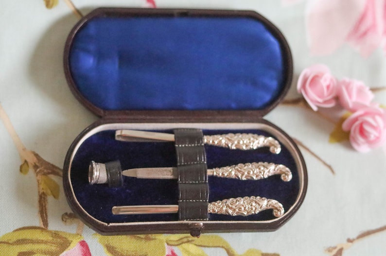 Puede incluir: Un juego de manicura vintage de plata con tres herramientas en un estuche forrado de terciopelo marr&oacute;n. Las herramientas tienen mangos de plata adornados y est&aacute;n dispuestas en el estuche.