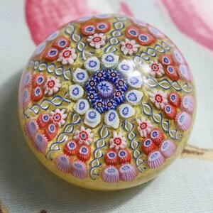 May include: A colorful glass paperweight with a swirling pattern of flowers in shades of pink, blue, and white. The paperweight is made of clear glass with a gold-colored background.