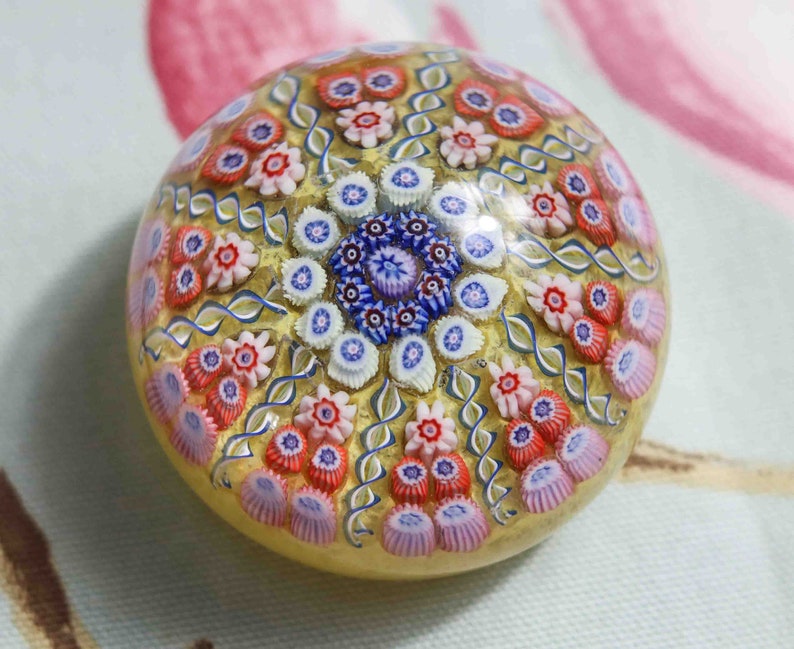 May include: A colorful glass paperweight with a swirling pattern of flowers in shades of pink, blue, and white. The paperweight is made of clear glass with a gold-colored background.