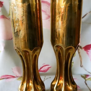 May include: Two gold-colored, vintage-style vases. The vases have a fluted base and a tall, slender body with etched floral designs. The vases are made of a shiny, reflective metal.