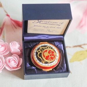 Puede incluir: Un estuche de terciopelo azul que contiene un compacto rojo y dorado con un diseño de esfera de reloj. El estuche tiene un certificado de autenticidad que dice "Dawn of a New...  This is number 16... Limited Edition of 1000"