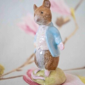 Peut inclure: Une petite figurine en porcelaine d'une souris portant une veste bleue et un gilet blanc. La souris est debout sur une base verte.