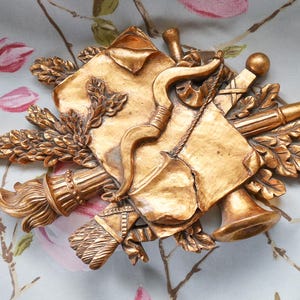 May include: A gold-coloured wall decoration with a bow, torch, horn, and other decorative elements. The design includes a central shield shape with a bow and arrow, surrounded by wheat and other embellishments. Set against a floral background.