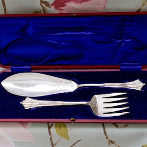 May include: A silver fish serving set, comprising a server and fork, displayed in a red-edged case with a blue velvet lining. The server has a broad, curved blade, and the fork has four prongs. Likely a vintage item.