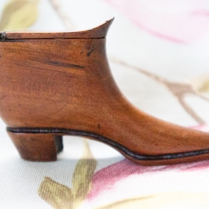 May include: A miniature wooden shoe, crafted from brown wood. The boot-shaped ornament features a pointed toe, a small heel, and a curved top. The wood grain is visible, giving texture to the decorative item. Likely a collectible or decorative piece.