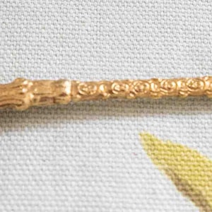 May include: A gold-colored spoon with ornate detailing. The spoon's bowl features embossed text and a crest. The handle is intricately designed, and the end of the handle has a decorative element.