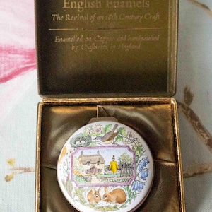 Puede incluir: Un estuche compacto redondo de esmalte blanco con una escena campestre, con una cabaña, conejos y una persona con un abrigo amarillo. El estuche compacto está en una caja dorada con el texto "English Enamels".