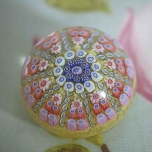 May include: A colorful glass paperweight with a swirling pattern of pink, white, blue, and yellow flowers. The paperweight is round and has a clear glass base.