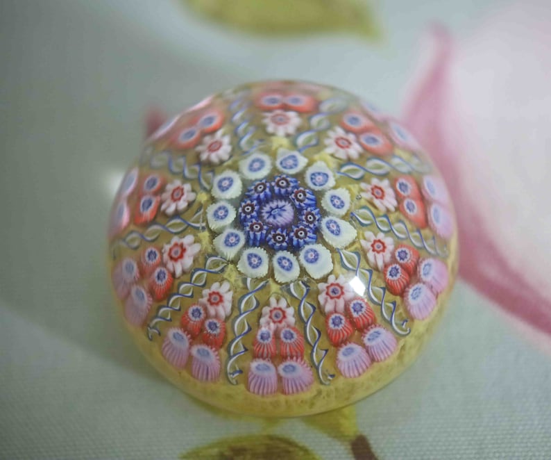 May include: A colorful glass paperweight with a swirling pattern of pink, white, blue, and yellow flowers. The paperweight is round and has a clear glass base.