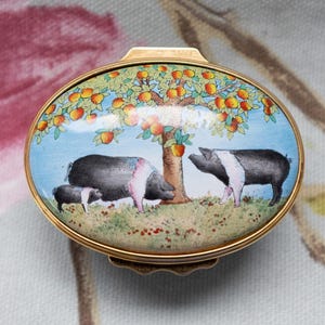 Pode incluir: Uma caixa de bugigangas oval dourada com uma tampa de esmalte pintada. A tampa apresenta uma cena pastoral com três porcos pretos e brancos sob uma macieira. O fundo é um céu azul claro.