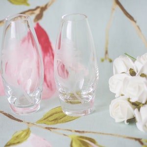 May include: Two clear glass vases with etched floral designs, sitting on a light blue and pink floral patterned surface. A bouquet of white roses is in the foreground.