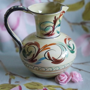 Può includere: Un'anfora in ceramica gialla con manico nero e disegni floreali dipinti in tonalità di blu, verde, arancio e marrone. L'anfora è appoggiata su un tessuto con motivo floreale.