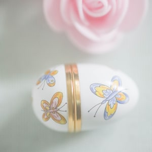 May include: A white ceramic egg-shaped trinket box with a gold hinged lid. The box is decorated with three colorful butterflies.