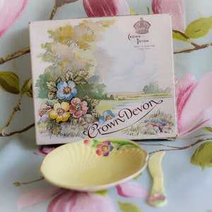 Könnte beinhalten: Ein Vintage-Buttergeschirr von Crown Devon mit einer gelben, muschelförmigen Schale und einem kleinen Buttermesser. Die Schachtel zeigt eine Aquarell-Illustration einer Landschaftsszene mit Blumen und dem Text "Crown Devon".