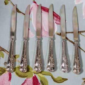 May include: Set of six silver butter knives with ornate handles. The knives have a polished finish and are arranged on a floral patterned tablecloth.