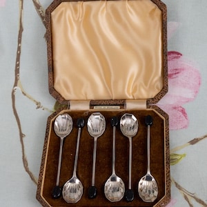 Könnte beinhalten: Ein Set aus sechs silbernen Löffeln mit schwarzen Akzenten, präsentiert in einer braunen, aufklappbaren Schatulle mit Satinfutter. Die Löffel haben facettierte Laffen und sind ordentlich in der Schatulle angeordnet.