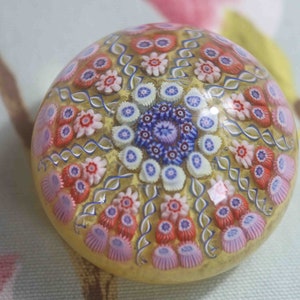 May include: A glass paperweight with a yellow background and a multi-colored floral pattern. The flowers are in shades of pink, red, blue, and white.
