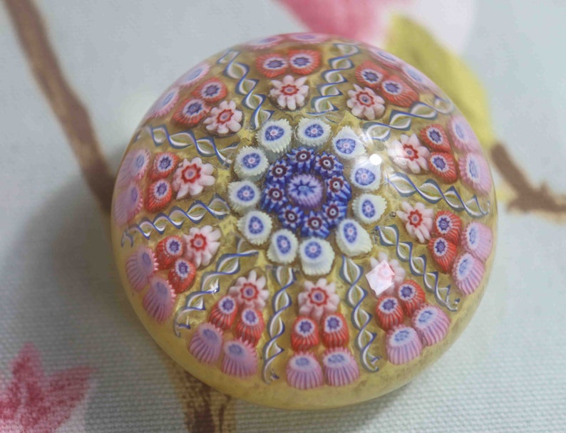 May include: A glass paperweight with a yellow background and a multi-colored floral pattern. The flowers are in shades of pink, red, blue, and white.