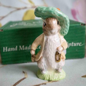 Może przedstawiać: Mała ceramiczna figurka Królika Piotrusia w zielonym kapeluszu i brązowej kurtce. Trzyma złoty klucz w prawej ręce. Figurka stoi na zielonej podstawie i jest otoczona zielonym pudełkiem z nadrukiem "Hand Made Sculpture".