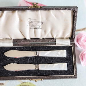 Könnte beinhalten: Zwei silberne Buttermesser mit Perlmuttgriffen in einer braunen, samtig ausgekleideten Schachtel. Die Schachtel trägt ein Etikett mit der Aufschrift "Thomas Armstrong & Brother Limited, 60 Deansgate, Manchester, England."
