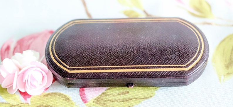 Puede incluir: Una caja ovalada de color marr&oacute;n oscuro con un borde dorado. La caja est&aacute; cerrada y tiene una superficie texturizada.