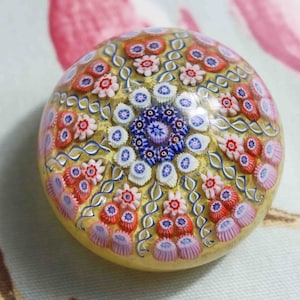 May include: A round glass paperweight with a gold background and a colorful floral pattern. The pattern features pink, red, blue, and white flowers.