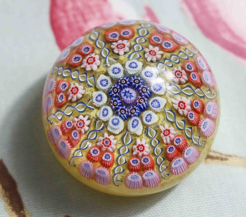 May include: A round glass paperweight with a gold background and a colorful floral pattern. The pattern features pink, red, blue, and white flowers.