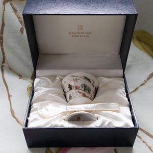 May include: A porcelain teacup and saucer set with a spoon, presented in a navy blue box. The teacup features a wildlife scene with elephants and other animals. The box is lined with white satin and has the words "Halcyon Days Porcelain" printed on the inside.