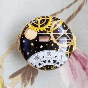 Puede incluir: Una caja de baratijas redonda con borde dorado y diseño celestial. La tapa negra presenta un sol amarillo, engranajes y estrellas. La mitad inferior tiene una esfera de reloj blanca con números romanos y símbolos de picas.