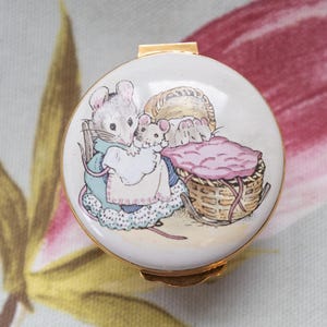 Może przedstawiać: Okrągłe, białe pudełko na drobiazgi ze złotą obwódką i zapięciem. Wieko zdobi urocza ilustracja dwóch myszy, z których jedna ma na sobie niebieską sukienkę i fartuch, z koszem i różowym kocem. Pudełko jest prawdopodobnie wykonane z porcelany lub emalii.