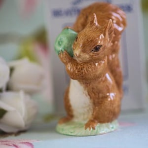Puede incluir: Una figurita de ardilla de cerámica marrón y blanca que sostiene una bellota verde. La ardilla está de pie sobre una base verde.
